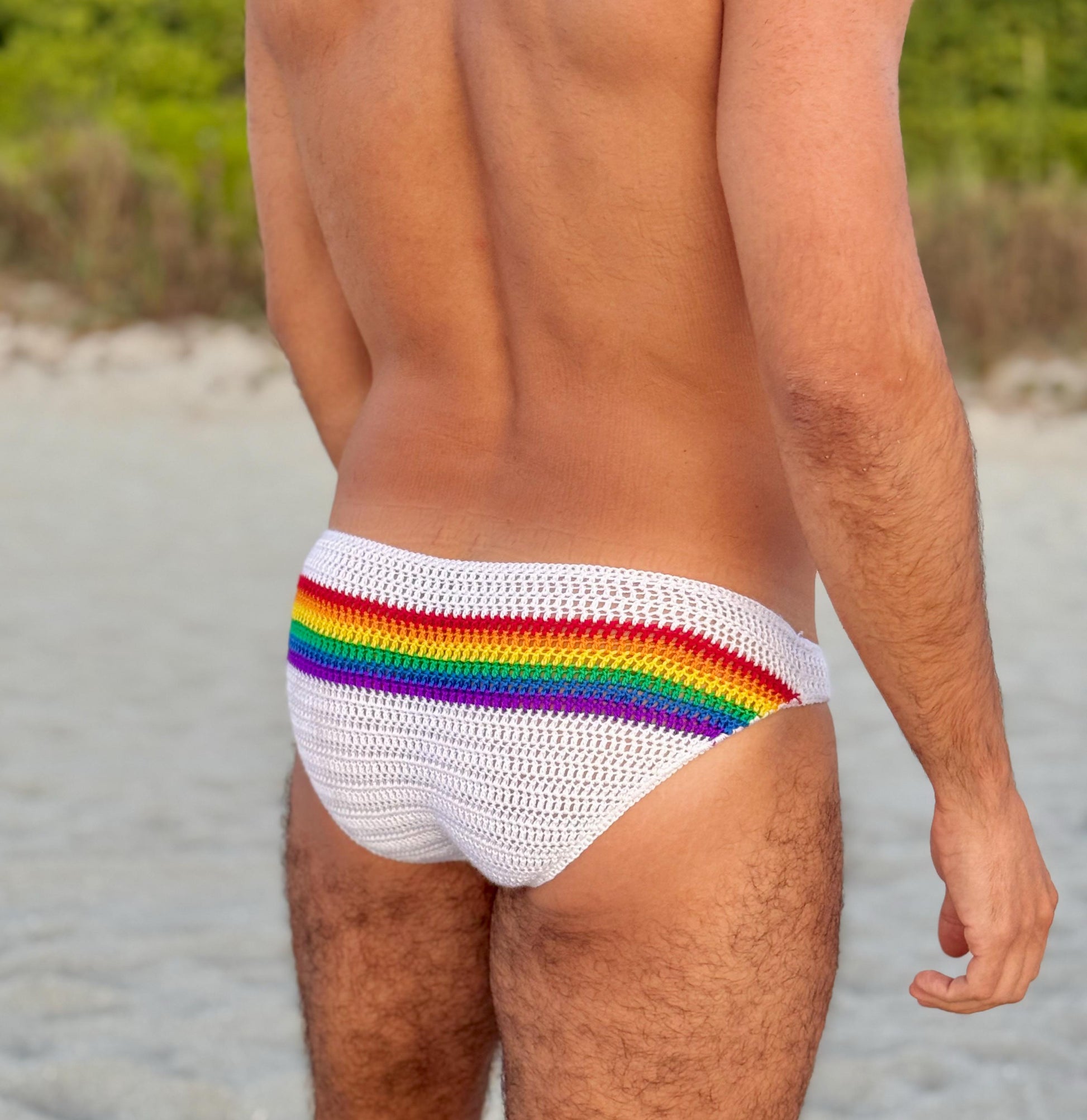 Crochet rainbow LGBTQ men’s briefs on a sandy background, beachwear aesthetic.
