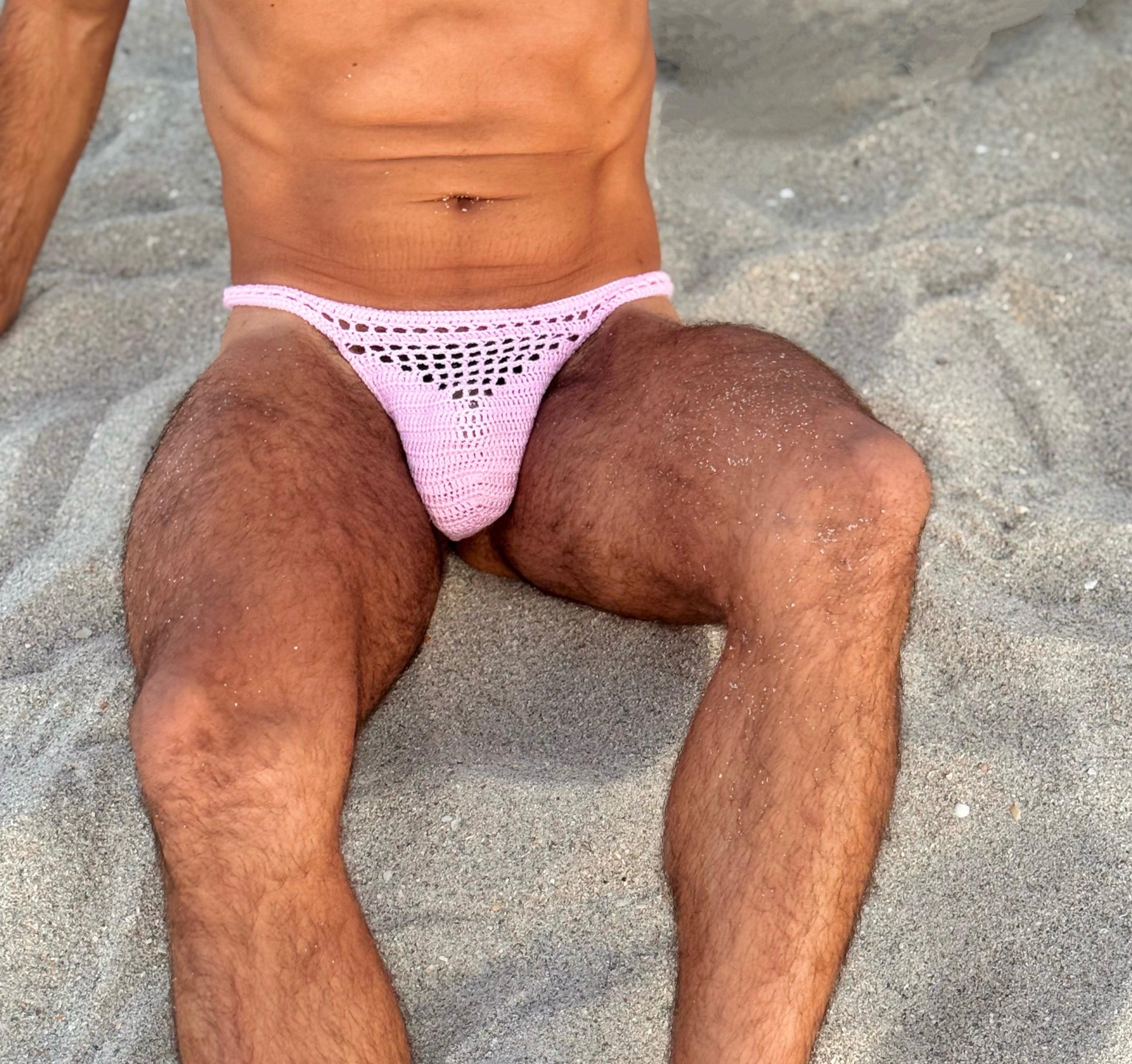 Crochet sheer men’s thong, displayed on a sand background.