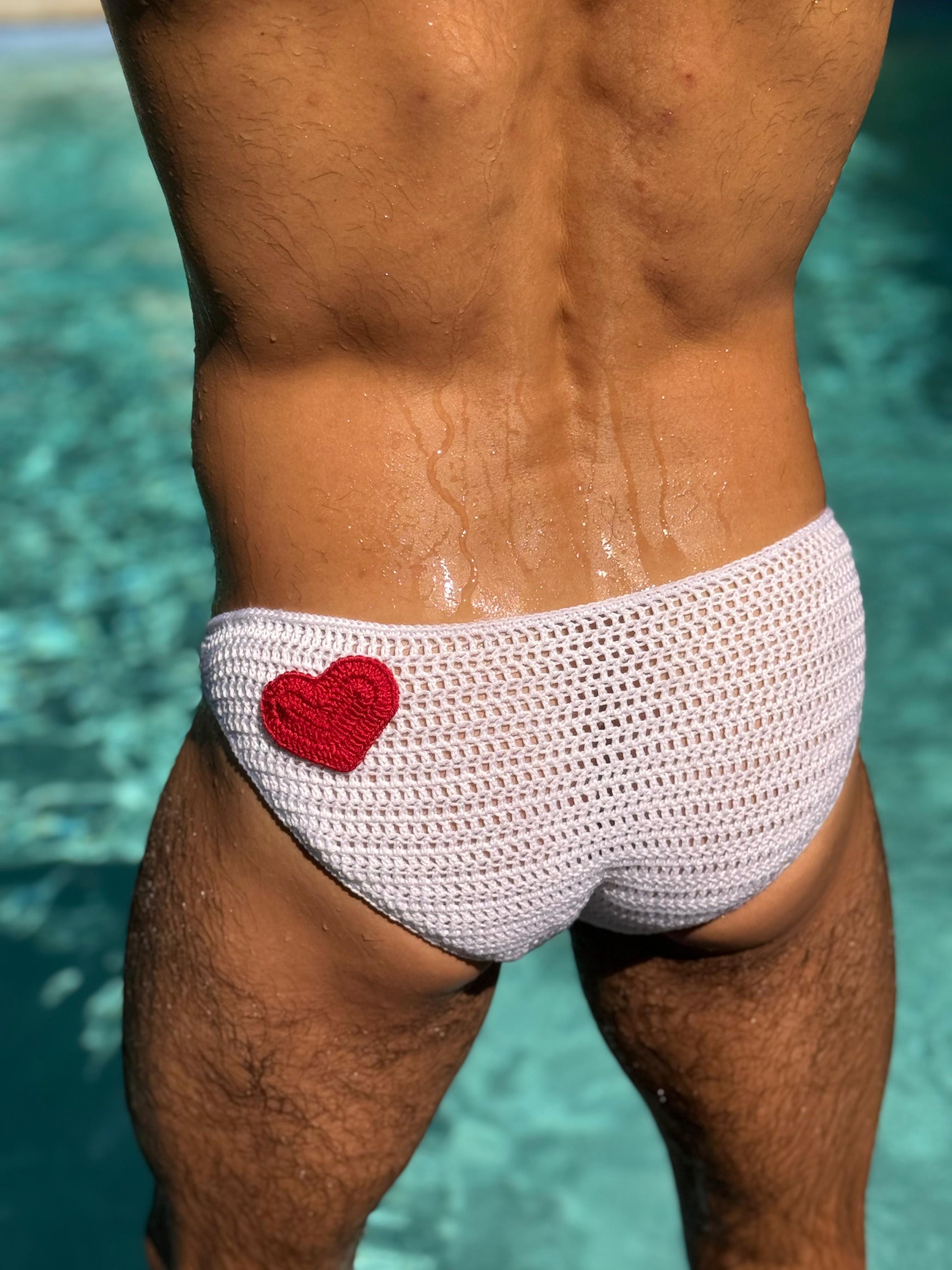 Close-up of crochet heart design on men's swim briefs, highlighting red heart on white crochet fabric.